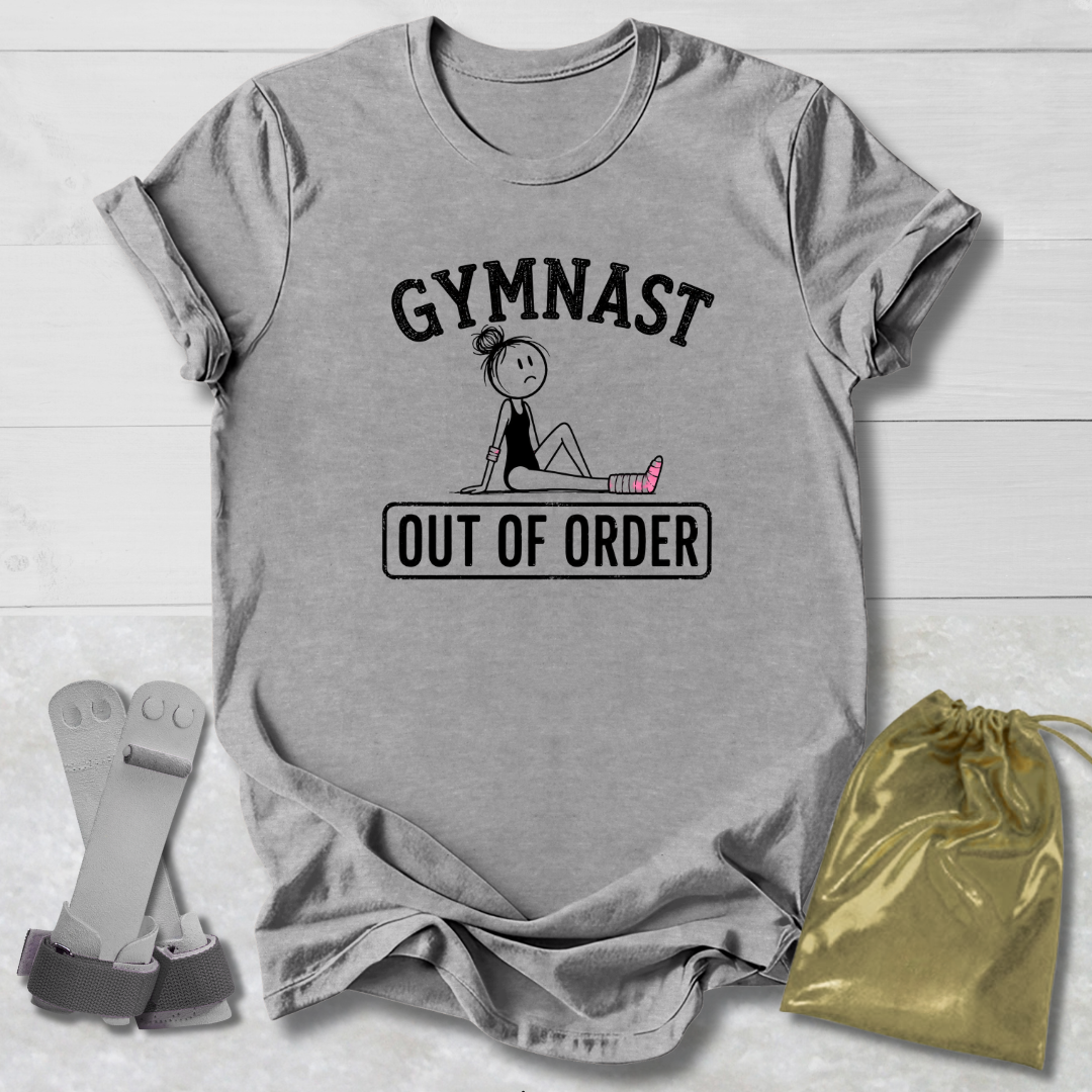A heather gray graphic t-shirt laid flat on a white wood background, featuring the text 'GYMNAST OUT OF ORDER' with a simple gymnast illustration. A gold drawstring bag and a pair of gray gymnast straps are placed beside the shirt.