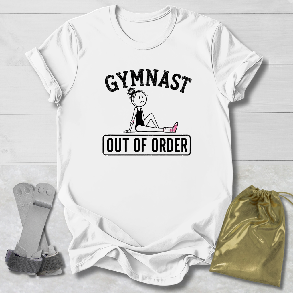 A white short-sleeve graphic T-shirt laid flat on a light wood background, featuring the words 'GYMNAST OUT OF ORDER' with a simple line drawing of a gymnast. A pair of gray straps and a gold drawstring bag are placed beside the shirt as styling props.