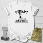 A white short-sleeve graphic T-shirt laid flat on a light wood background, featuring the words 'GYMNAST OUT OF ORDER' with a simple line drawing of a gymnast. A pair of gray straps and a gold drawstring bag are placed beside the shirt as styling props.