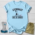 A light blue short-sleeve t-shirt laid flat on a white wood background, featuring a black graphic of a gymnast and the words 'GYMNAST OUT OF ORDER.' Two gray and blue gymnastic straps and a shiny gold drawstring bag are placed beside the shirt as styling props.