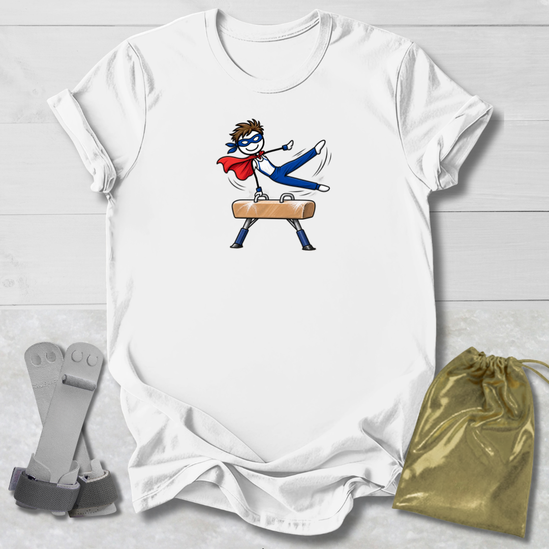 A white short-sleeve T-shirt laid flat on a light wood background, featuring a cartoon graphic of a superhero in a blue mask and red cape riding a pommel horse. A gold drawstring pouch and gray straps are placed beside the shirt.