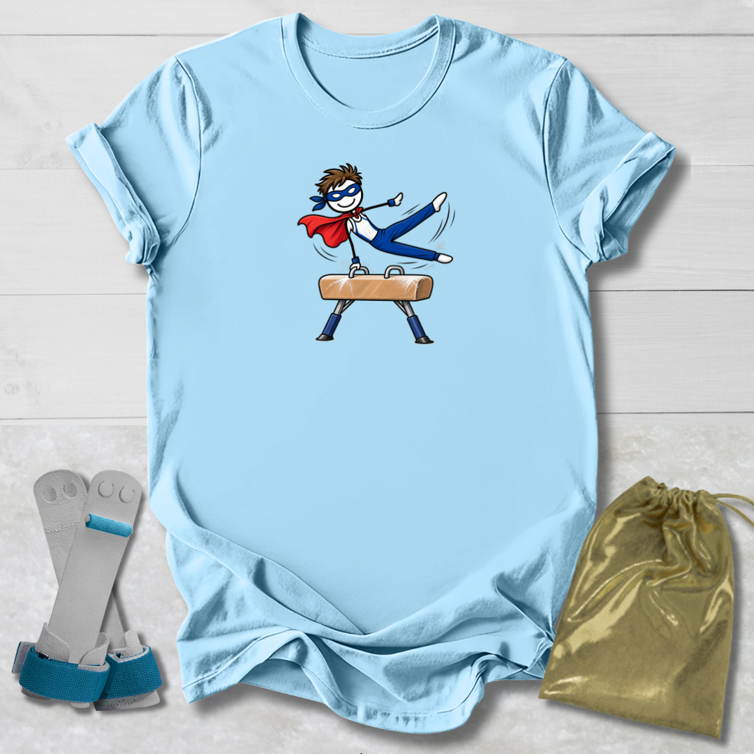 A light blue short-sleeve T-shirt laid flat on a white wood background, featuring a cartoon graphic of a superhero in a red cape and blue mask performing a kick on a pommel horse. A gold drawstring bag and a pair of blue-and-gray leg straps are placed beside the shirt as included accessories.