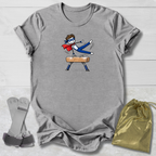 A heather gray short-sleeve t-shirt with a cartoon graphic of a gymnast in a red cape on a balance beam, styled flat-lay with a gold drawstring bag and a pair of gray straps on a white wood background.