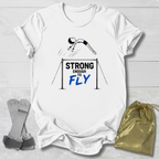 A white graphic t-shirt laid flat on a light wood background, featuring a gymnast illustration and the text 'STRONG ENOUGH TO FLY' in black and blue. A gray climbing harness and a gold drawstring pouch are placed beside the shirt as styling props.