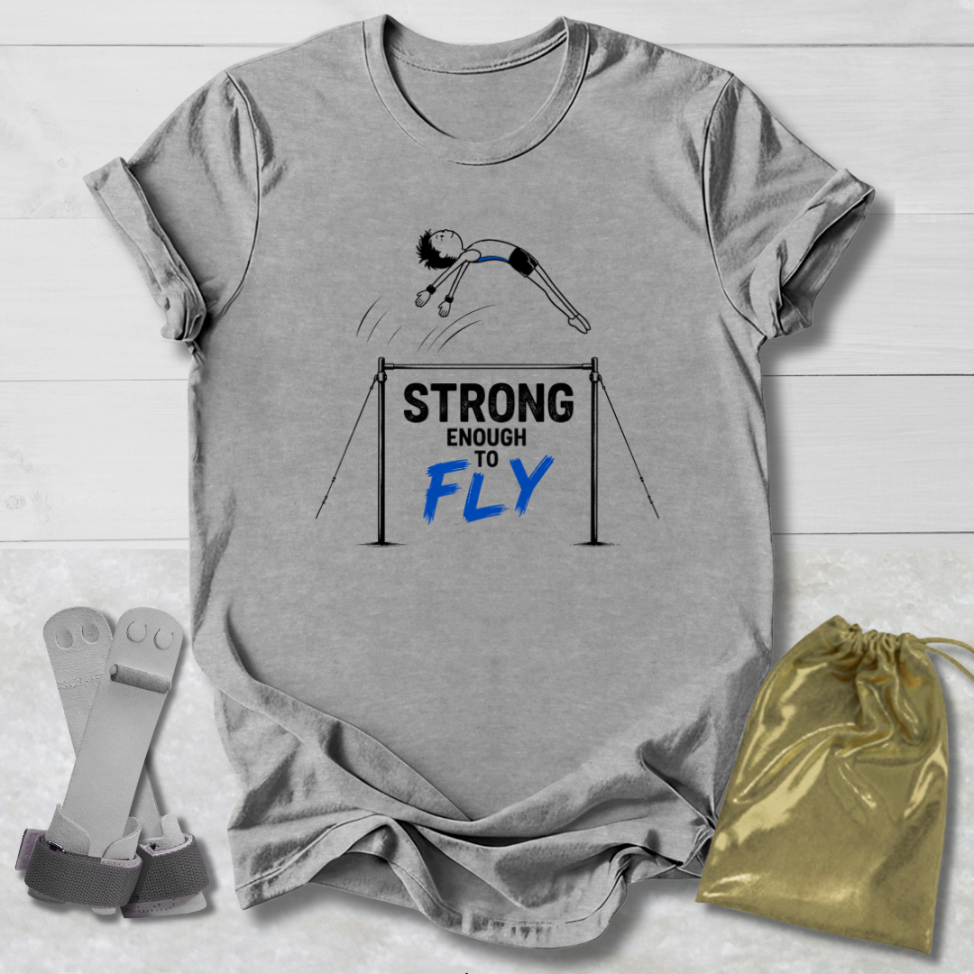 A heather gray graphic T-shirt laid flat on a white wood background, featuring a gymnast illustration and the text 'STRONG ENOUGH TO FLY' in black and blue. A gold drawstring pouch and a pair of gray straps are placed beside the shirt as styling props.