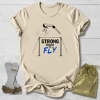 A beige graphic T-shirt laid flat on a white wood background, featuring a gymnast illustration and the text 'STRONG ENOUGH TO FLY' in black and blue. A gold drawstring bag and gymnastics straps are placed beside the shirt as styling props.
