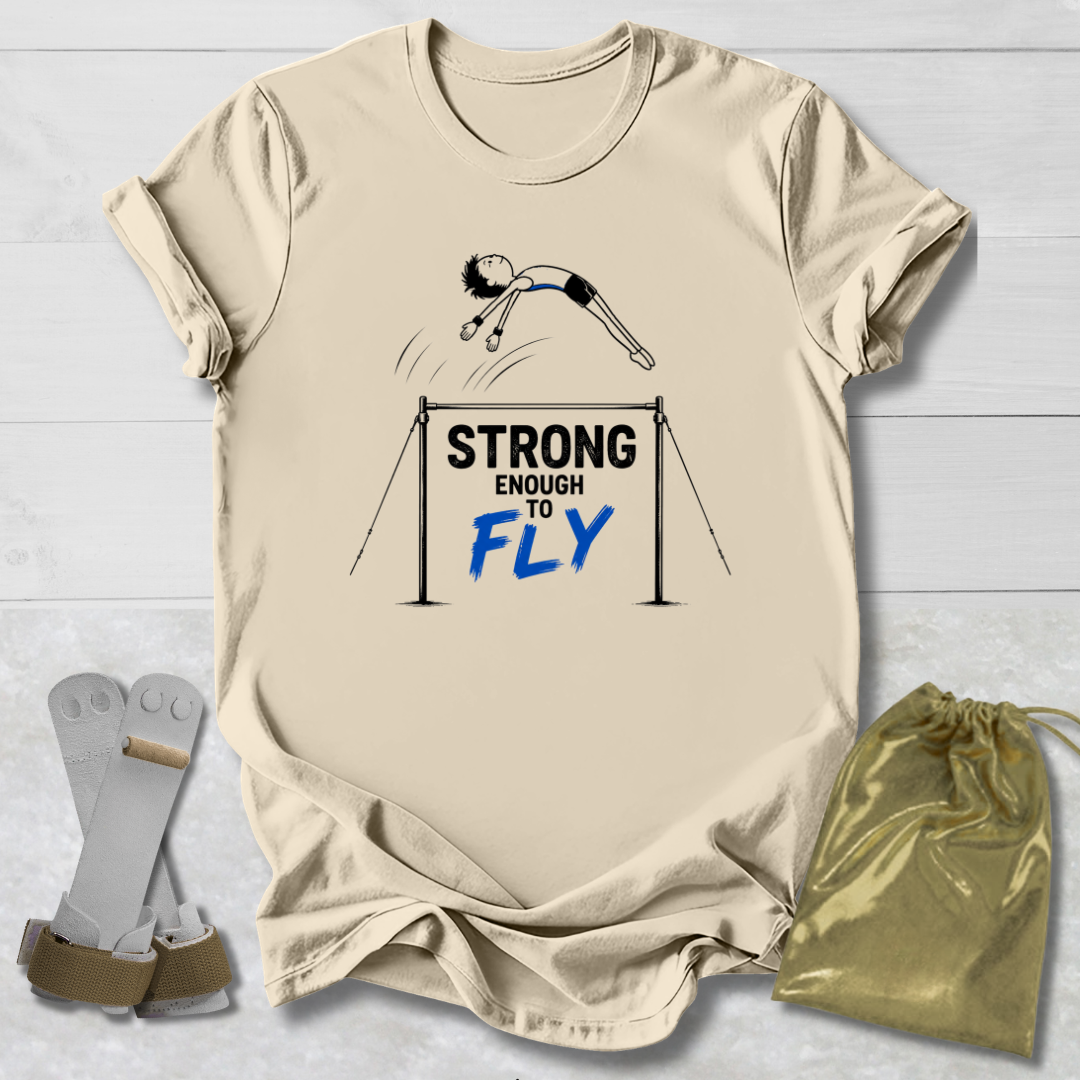A beige graphic T-shirt laid flat on a white wood background, featuring a gymnast illustration and the text 'STRONG ENOUGH TO FLY' in black and blue. A gold drawstring bag and gymnastics straps are placed beside the shirt as styling props.
