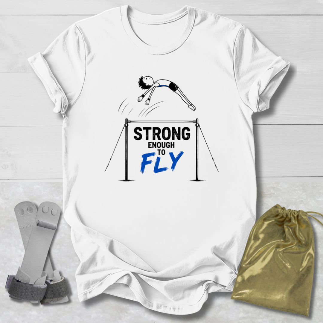 A white graphic t-shirt laid flat on a light wood background, featuring a gymnast illustration and the text 'STRONG ENOUGH TO FLY' in black and blue. A gray climbing harness and a gold drawstring pouch are placed beside the shirt as styling props.