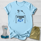 A light blue graphic T-shirt laid flat on a white wood background, featuring a gymnast illustration and the text 'STRONG ENOUGH TO FLY.' Two athletic support sleeves and a gold drawstring bag are placed beside the shirt as styling props.
