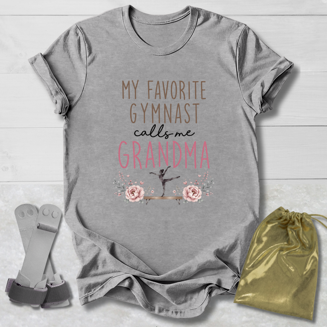 Gray short-sleeve T-shirt laid flat on a light wood background, printed with the phrase 'MY FAVORITE GYMNAST calls me GRANDMA' in brown, black, and pink lettering, with a small gymnast silhouette and floral graphic. A pair of gray resistance bands and a gold drawstring bag are placed beside the shirt as styling props.