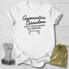 A white short-sleeve graphic T-shirt laid flat on a light wood background, with black text reading 'Gymnastics Grandma: like a normal grandma but COOLER.' and a bench illustration.