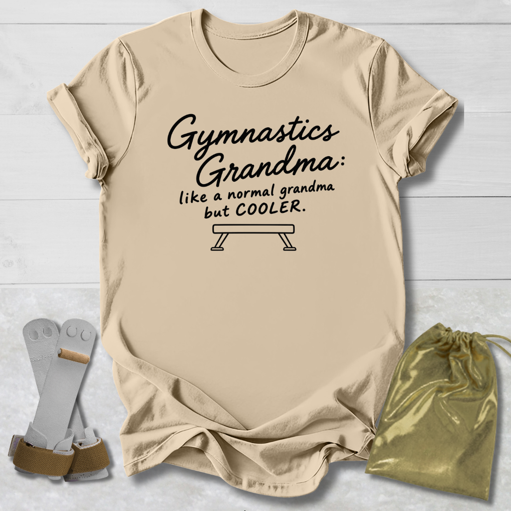 A beige graphic t-shirt laid flat on a white wood background, featuring the black text 'Gymnastics Grandma: like a normal grandma but COOLER.' with a bench graphic.