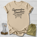 A beige graphic t-shirt laid flat on a white wood background, featuring the black text 'Gymnastics Grandma: like a normal grandma but COOLER.' with a bench graphic.