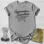 A light gray graphic t-shirt laid flat on a white wood background, featuring the text 'Gymnastics Grandma: like a normal grandma but COOLER.' with a bench graphic.