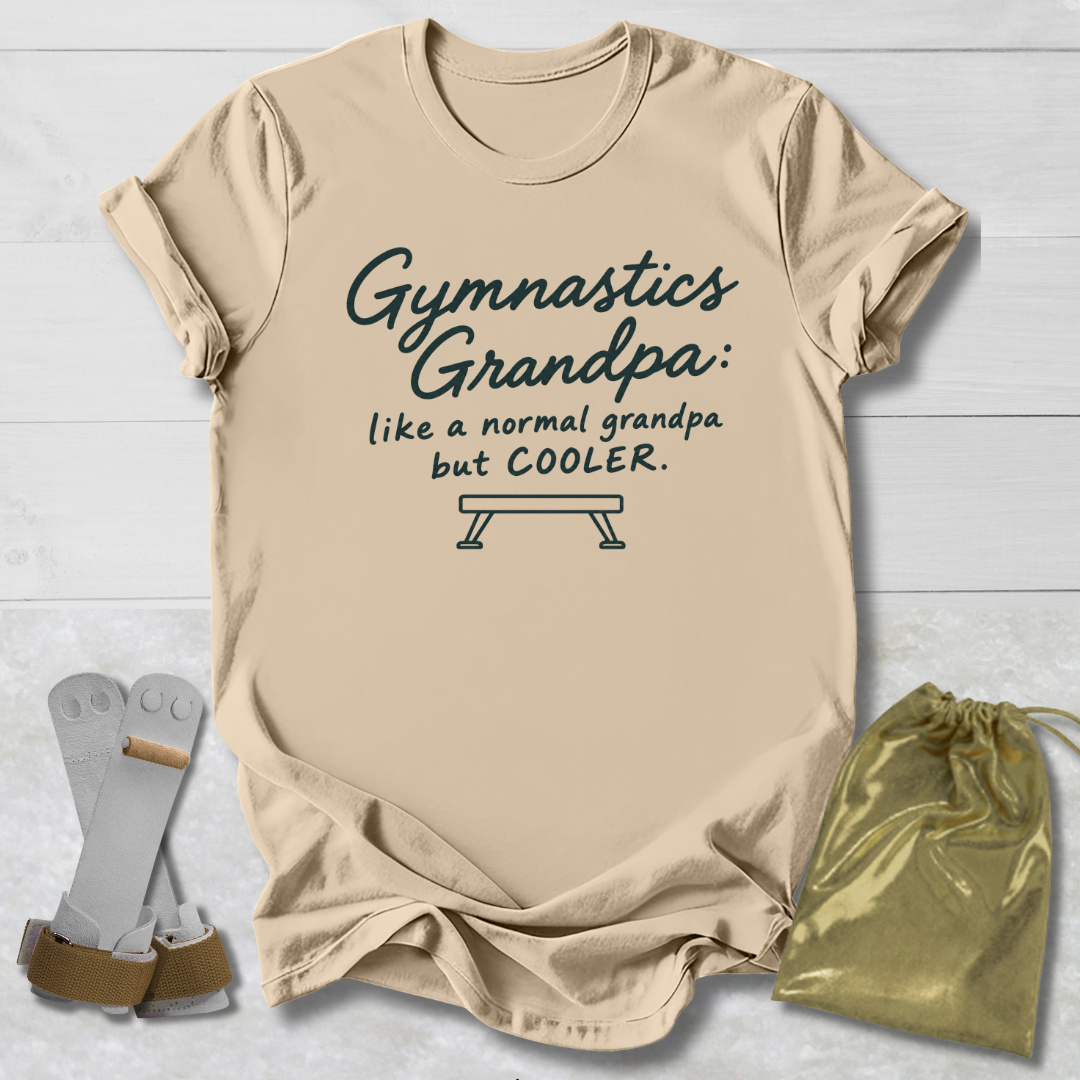 A beige graphic t-shirt laid flat on a white wood background, featuring the text 'Gymnastics Grandpa: like a normal grandpa but COOLER.' with a bench graphic.