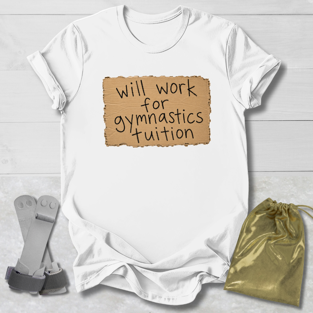 A white short-sleeve T-shirt laid flat on a light wood background, featuring a tan cardboard-style graphic with the handwritten text 'will work for gymnastics tuition.' A gray strap and a shiny gold drawstring pouch are placed beside the shirt as styling props.