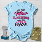 A light blue graphic T-shirt laid flat on a white wood background, with the text 'I'M ONE New FLOOR ROUTINE AWAY FROM LIVING IN MY CAR.' in pink, gray, and black lettering.