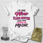 A white graphic T-shirt laid flat on a light wood background, with gray and pink text reading, "I'M ONE New FLOOR ROUTINE AWAY FROM LIVING IN MY CAR." Two gray straps and a small gold drawstring pouch are placed beside the shirt as styling props.