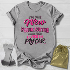 A heather gray graphic T-shirt laid flat on a white wood background, with a metallic pink and black slogan reading “I’m One New Floor Routine Away From Living In My Car.” A small gold drawstring pouch and gray strap accessories are placed beside the shirt.