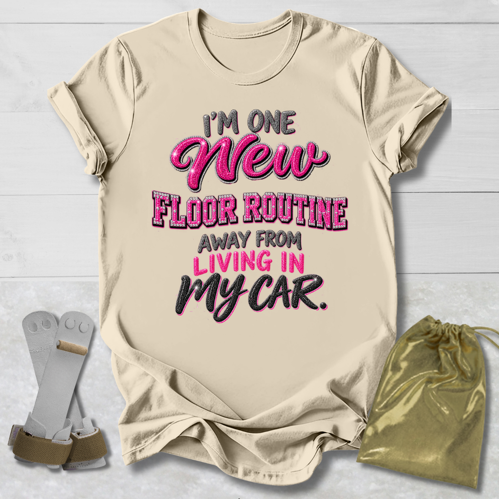 A beige graphic t-shirt laid flat on a white wood background, featuring the text 'I'M ONE New FLOOR ROUTINE AWAY FROM LIVING IN MY CAR.' in pink and black.
