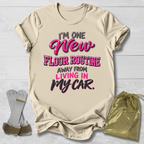 A beige graphic t-shirt laid flat on a white wood background, featuring the text 'I'M ONE New FLOOR ROUTINE AWAY FROM LIVING IN MY CAR.' in pink and black.