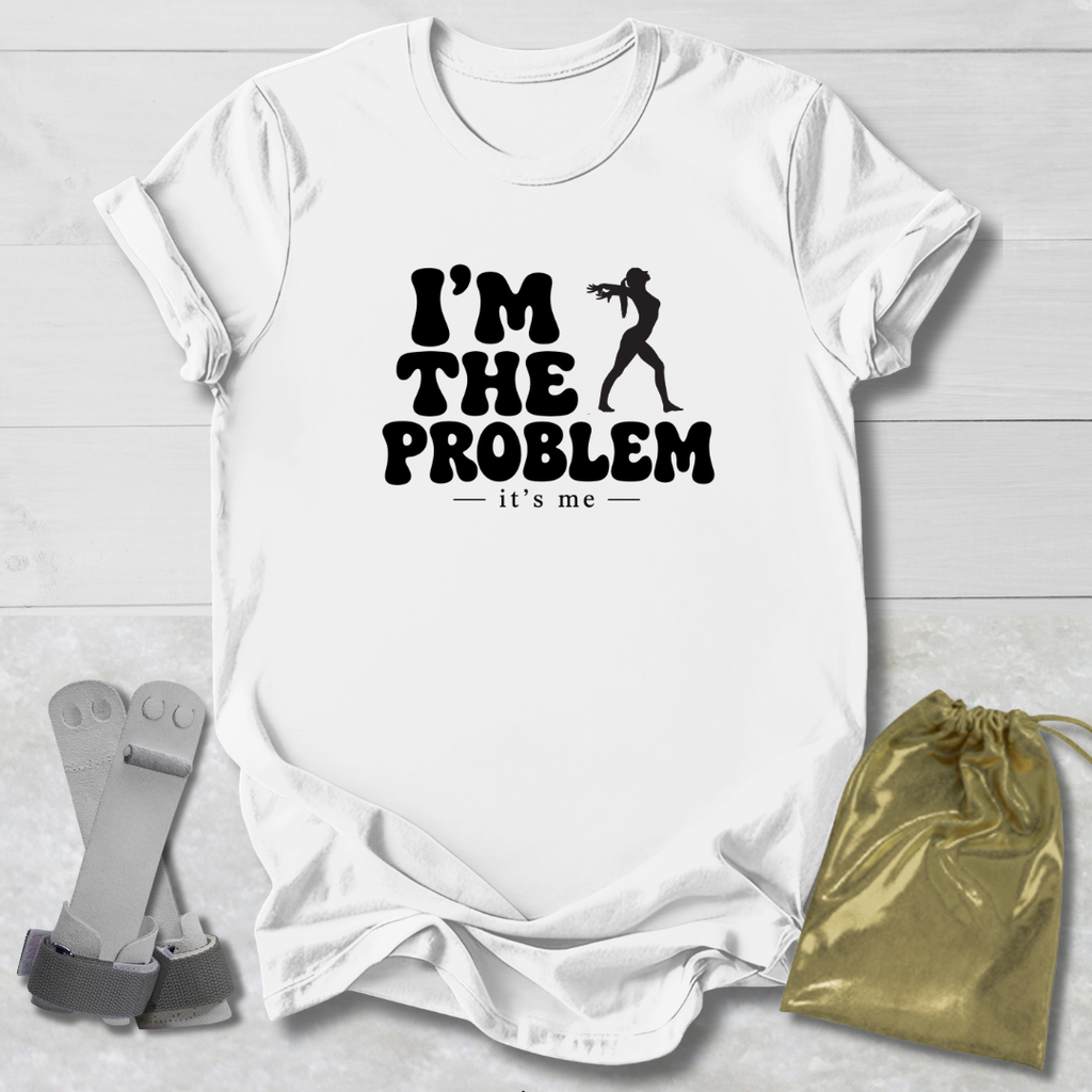 A white short-sleeve graphic T-shirt laid flat on a light wood background, with black text reading “I’M THE PROBLEM it’s me” and a black silhouette of a person holding a gun. A metallic gold drawstring pouch and a gray strap-like accessory are placed beside the shirt as styling props.