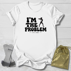 A white short-sleeve graphic T-shirt laid flat on a light wood background, with black text reading “I’M THE PROBLEM it’s me” and a black silhouette of a person holding a gun. A metallic gold drawstring pouch and a gray strap-like accessory are placed beside the shirt as styling props.