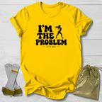 A bright yellow graphic t-shirt laid flat on a white wood background, featuring the black text 'I'M THE PROBLEM it's me' with a silhouette of a person holding a gun. A gold drawstring pouch and a pair of white and gold accessories are placed beside the shirt.