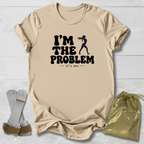 A beige graphic t-shirt laid flat on a white wood background, featuring the black text 'I'M THE PROBLEM it's me' with a silhouette of a person holding a gun. A gold drawstring pouch and a pair of gray and brown gloves are placed beside the shirt as props.