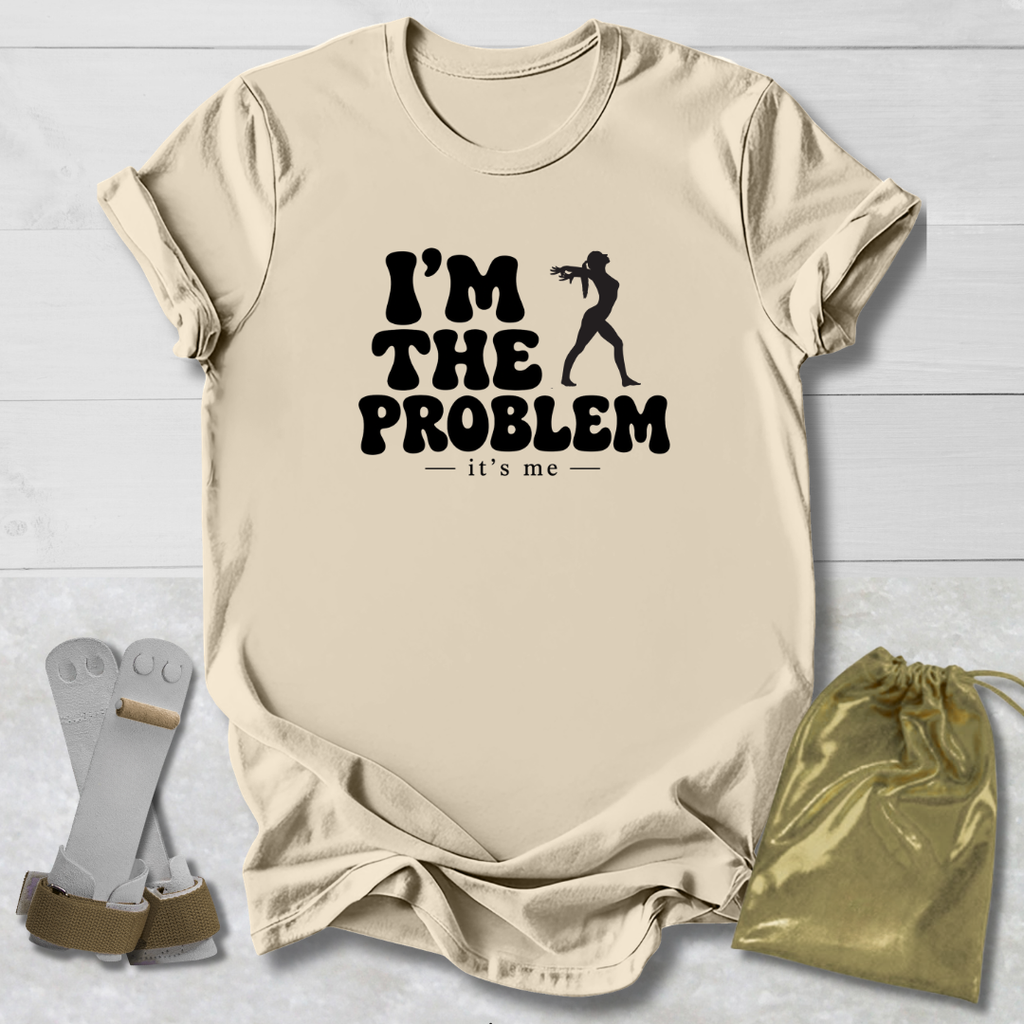 A beige graphic t-shirt laid flat on a white wood background, featuring the black text 'I'M THE PROBLEM it's me' with a silhouette of a person holding a gun. A pair of white and brown boxing gloves and a gold drawstring pouch are placed beside the shirt as props.