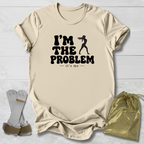 A beige graphic t-shirt laid flat on a white wood background, featuring the black text 'I'M THE PROBLEM it's me' with a silhouette of a person holding a gun. A pair of white and brown boxing gloves and a gold drawstring pouch are placed beside the shirt as props.