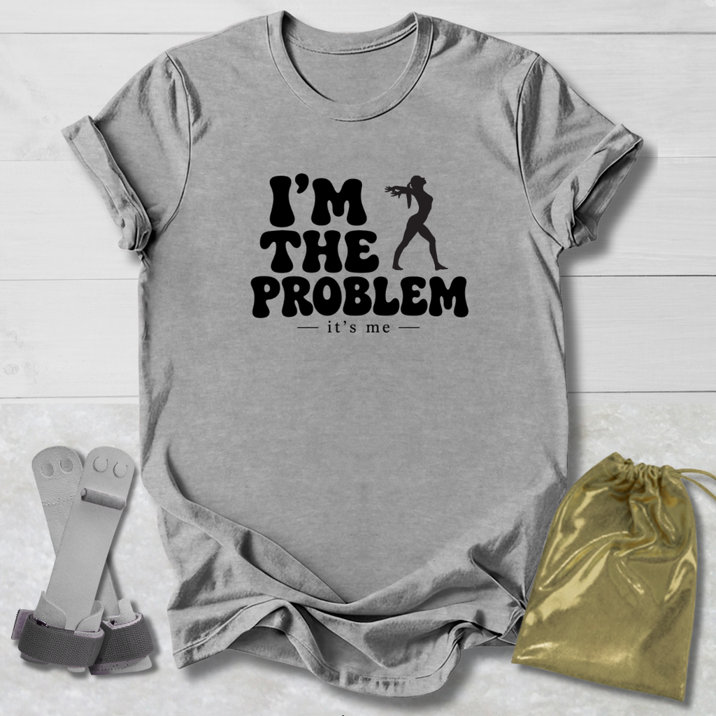 A heather gray short-sleeve t-shirt with a black graphic print reading 'I'M THE PROBLEM it's me' alongside a silhouette of a person holding a gun, shown in a flat-lay arrangement with a gold drawstring pouch and gray straps on a white wood-plank background.
