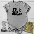A heather gray short-sleeve t-shirt with a black graphic print reading 'I'M THE PROBLEM it's me' alongside a silhouette of a person holding a gun, shown in a flat-lay arrangement with a gold drawstring pouch and gray straps on a white wood-plank background.
