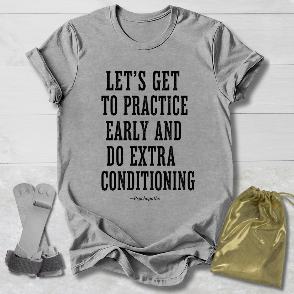 A heather gray graphic T-shirt laid flat on a white wood background, with black text reading “LET’S GET TO PRACTICE EARLY AND DO EXTRA CONDITIONING” and a small “-Psychopaths” signature underneath. A gold drawstring pouch and a gray strap accessory are placed beside the shirt as styling props.