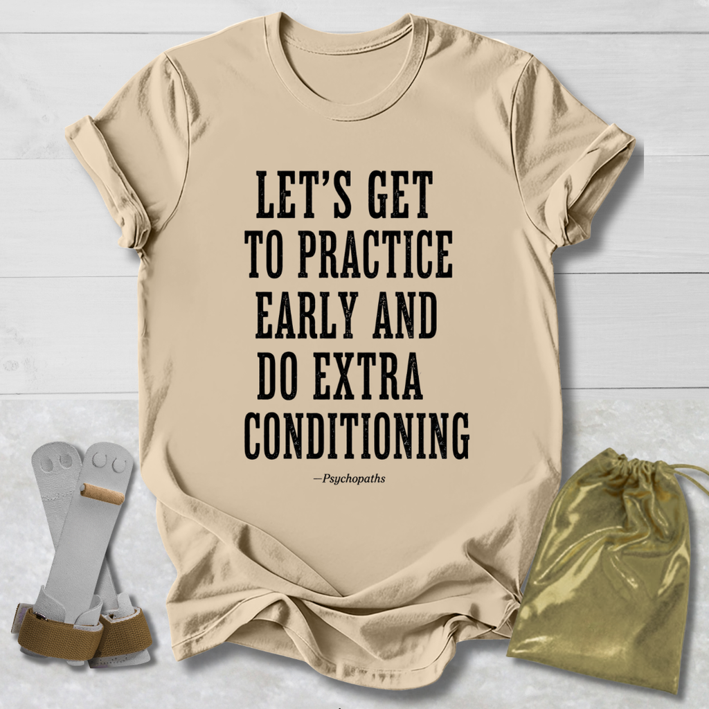 A beige graphic T-shirt laid flat on a white wood background, with black text reading “LET’S GET TO PRACTICE EARLY AND DO EXTRA CONDITIONING” and a small “-Psychopaths” attribution below. A pair of white and tan shin guards and a shiny gold drawstring pouch are placed around the shirt as styling props.