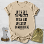 A beige graphic T-shirt laid flat on a white wood background, with black text reading “LET’S GET TO PRACTICE EARLY AND DO EXTRA CONDITIONING” and a small “-Psychopaths” attribution below. A pair of white and tan shin guards and a shiny gold drawstring pouch are placed around the shirt as styling props.