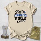 A beige graphic t-shirt laid flat on a white wood background, featuring the text 'Best GYMNASTICS UNCLE Ever!' in black and blue. A gold drawstring bag and a pair of white and gold gymnastics straps are placed beside the shirt.