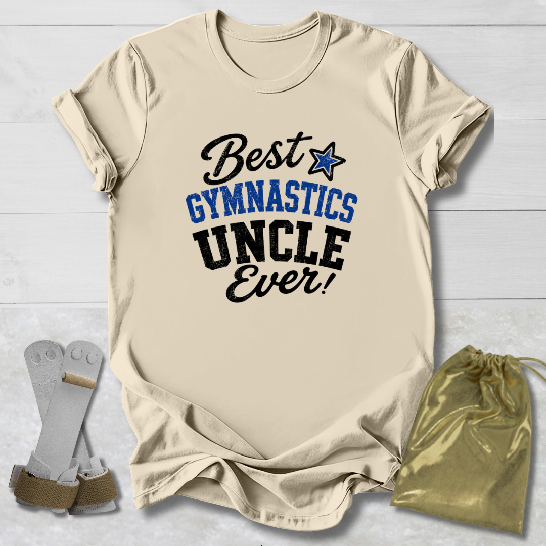 A beige graphic t-shirt laid flat on a white wood background, featuring the text 'Best GYMNASTICS UNCLE Ever!' in black and blue. A gold drawstring bag and a pair of white and gold gymnastics straps are placed beside the shirt.