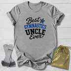 A heather gray graphic t-shirt laid flat on a white wood background, featuring the text 'Best GYMNASTICS UNCLE Ever!' in black and blue. A gold drawstring bag and a pair of gray wristbands are placed beside the shirt.
