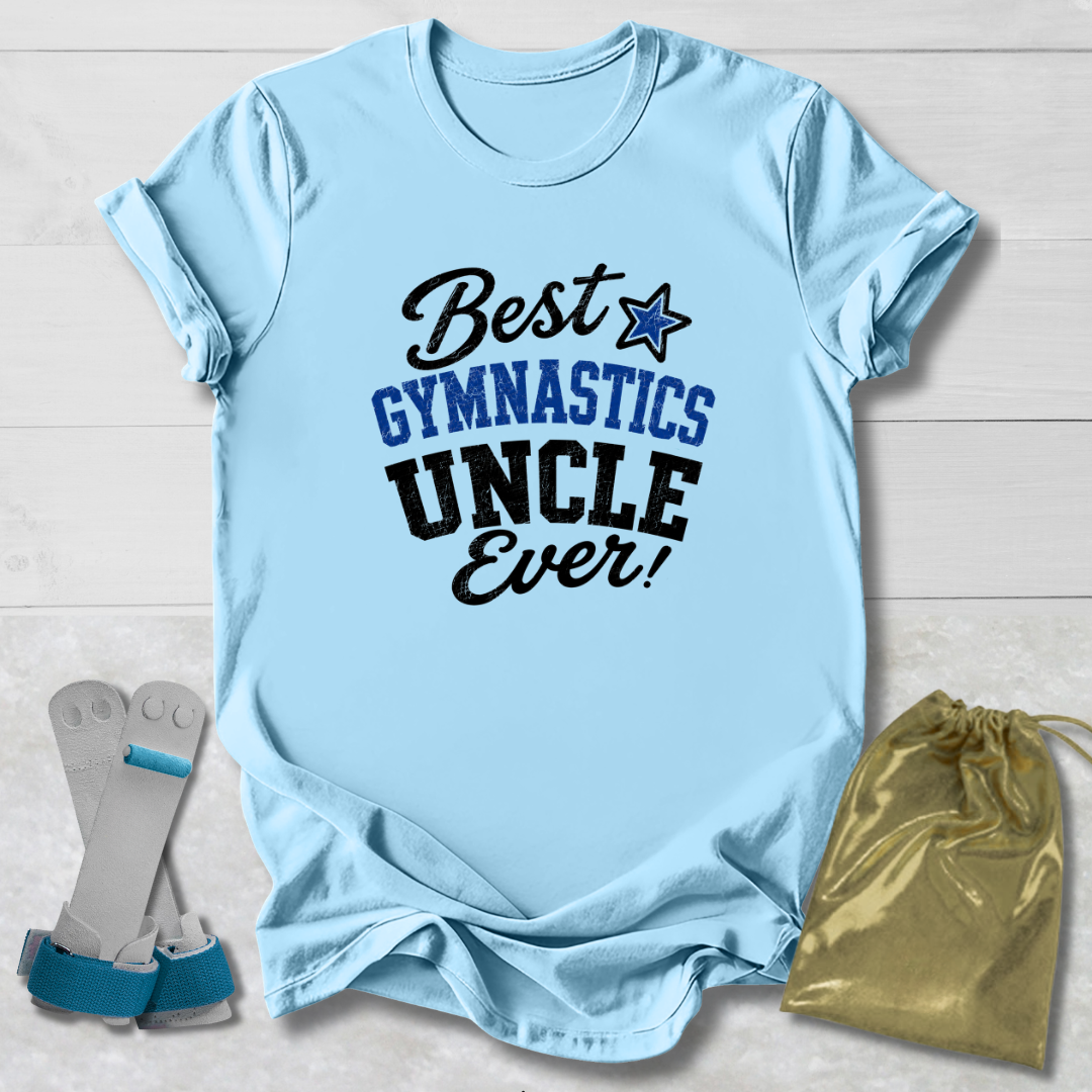 A light blue short-sleeve t-shirt laid flat on a white wood background, printed with the phrase 'Best GYMNASTICS UNCLE Ever!' in black and blue lettering with a blue star. A pair of gymnastics trunks and a gold drawstring pouch are placed beside the shirt as styling props.