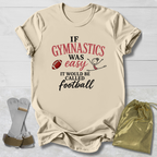 A beige graphic t-shirt laid flat on a white wood background, featuring the text 'IF GYMNASTICS WAS easy IT WOULD BE CALLED football' in black and red lettering with a small football and gymnast illustration. A gold drawstring pouch and a pair of gymnastics straps are placed beside the shirt.