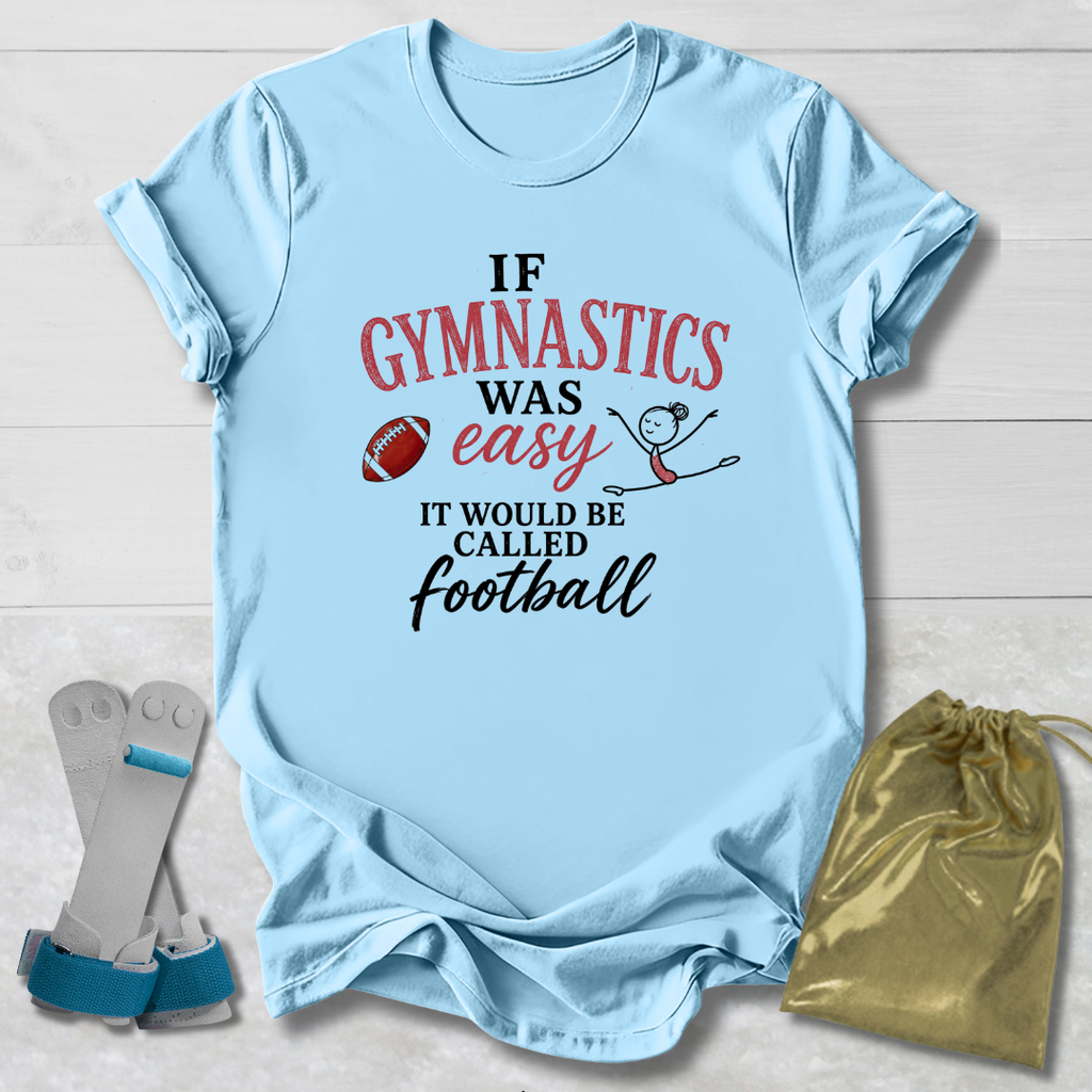 A light blue graphic t-shirt laid flat on a white wood background, featuring the text 'IF GYMNASTICS WAS easy IT WOULD BE CALLED football' in red, pink, and black lettering, accompanied by a small football and stick figure drawing. A gold drawstring bag and a pair of gray and blue gymnastics shoes are placed beside the shirt.