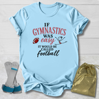 A light blue graphic t-shirt laid flat on a white wood background, featuring the text 'IF GYMNASTICS WAS easy IT WOULD BE CALLED football' in red, pink, and black lettering, accompanied by a small football and stick figure drawing. A gold drawstring bag and a pair of gray and blue gymnastics shoes are placed beside the shirt.
