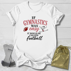 A white short-sleeve graphic T-shirt laid flat on a light wood background, with a gymnastics-themed slogan printed in red and black text and small football and stick-figure graphics. A gold drawstring pouch and gray straps are placed beside the shirt as styling props.