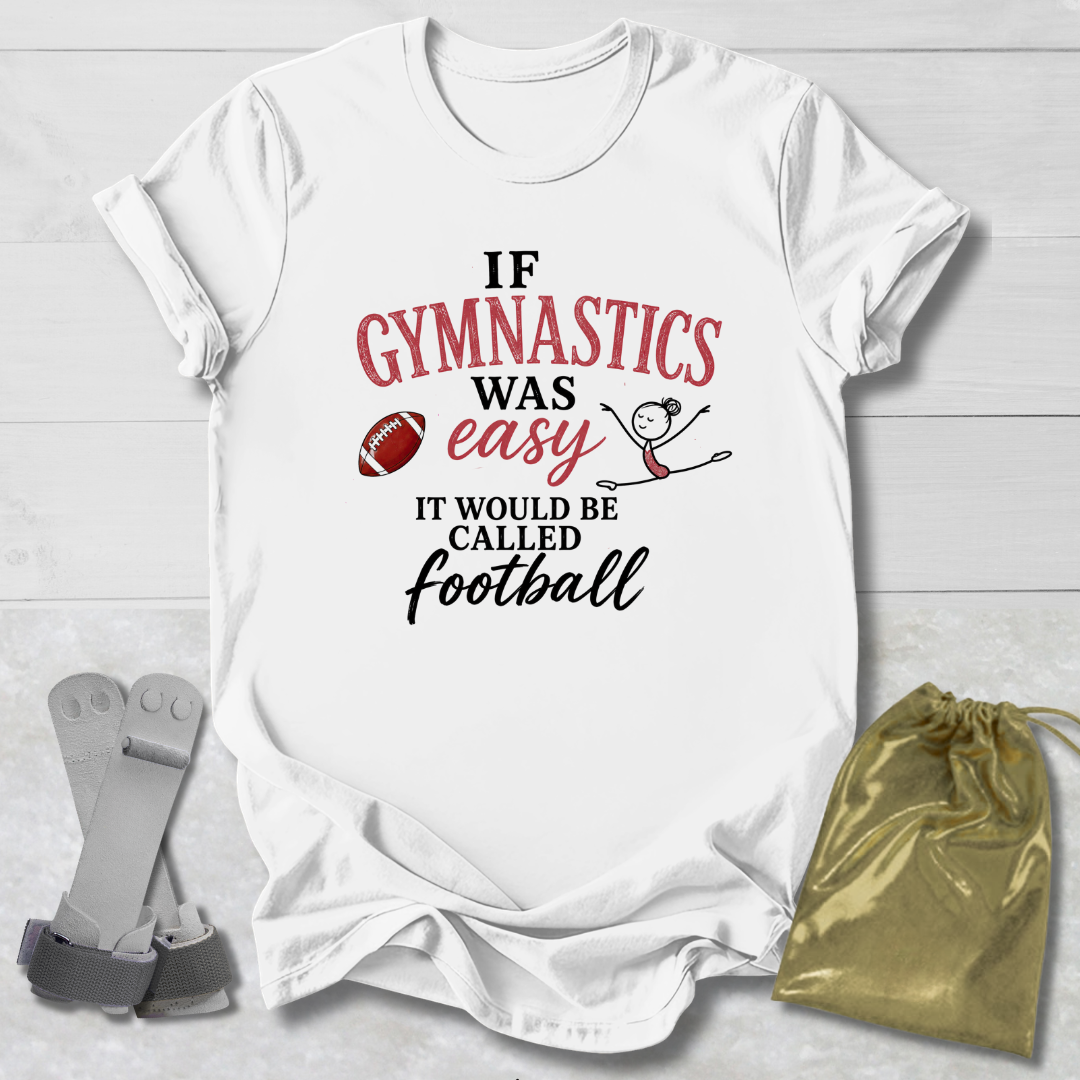 A white short-sleeve graphic T-shirt laid flat on a light wood background, with a gymnastics-themed slogan printed in red and black text and small football and stick-figure graphics. A gold drawstring pouch and gray straps are placed beside the shirt as styling props.