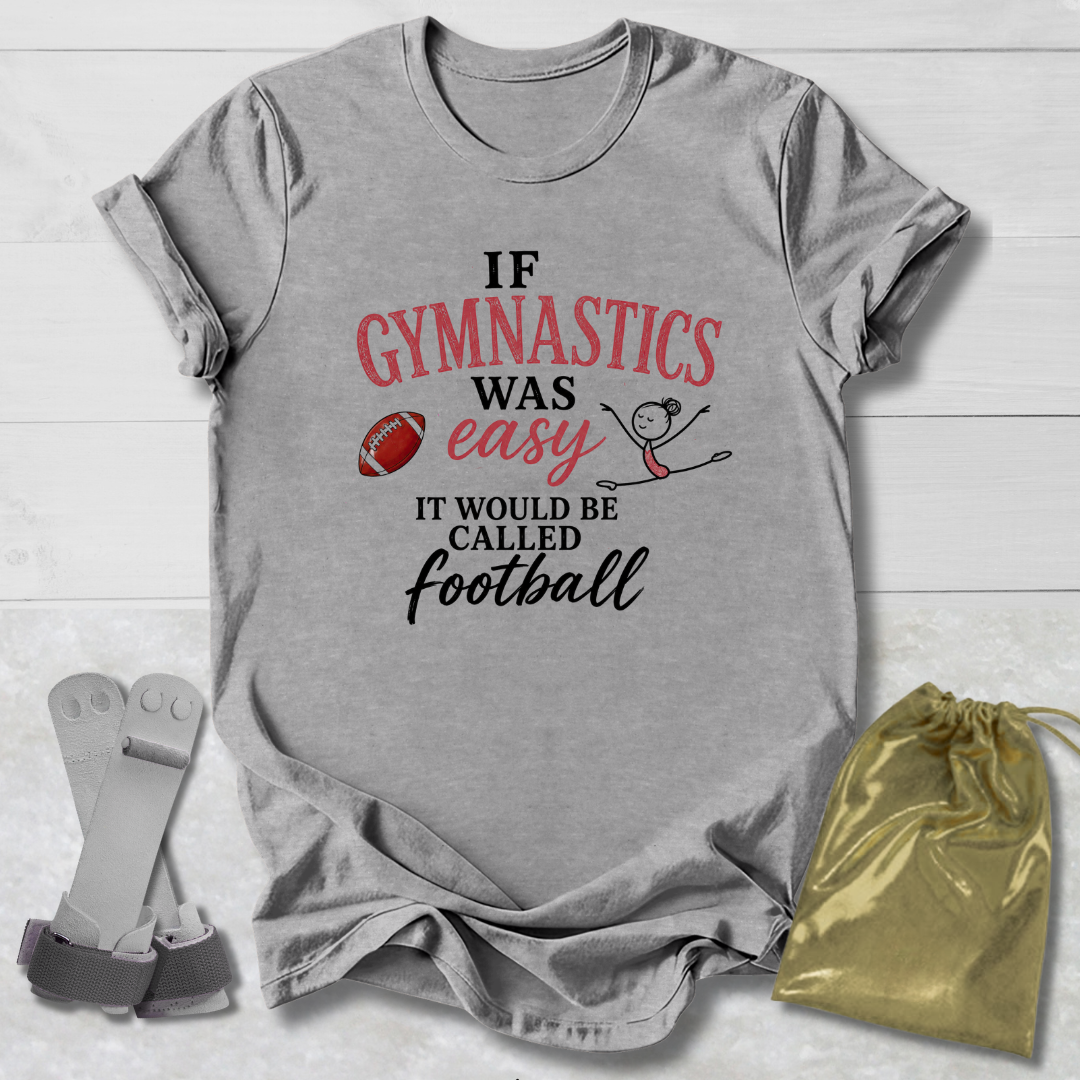 A heather gray graphic t-shirt laid flat on a white wood background, featuring the text 'IF GYMNASTICS WAS easy IT WOULD BE CALLED football' in black and red lettering with a small football and stick-figure graphic. A gold drawstring bag and a pair of gray athletic straps are placed beside the shirt.