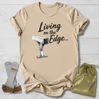 Beige balance beam graphic T-shirt laid flat on a white wood background, printed with the words 'Living on the Edge.