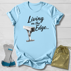 A light blue graphic T-shirt laid flat on a white wood background, printed with the words 'Living on the Edge.