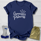Navy short-sleeve t-shirt with 'Gymnastics Princess' printed in white script under a small crown graphic, shown on a light wood background with a pair of gymnastics trunks and a gold drawstring bag.
