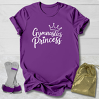 A purple short-sleeve t-shirt with 'Gymnastics Princess' printed in white script under a small crown graphic, shown on a light wood background with a pair of gymnastics trunks and a gold drawstring bag.